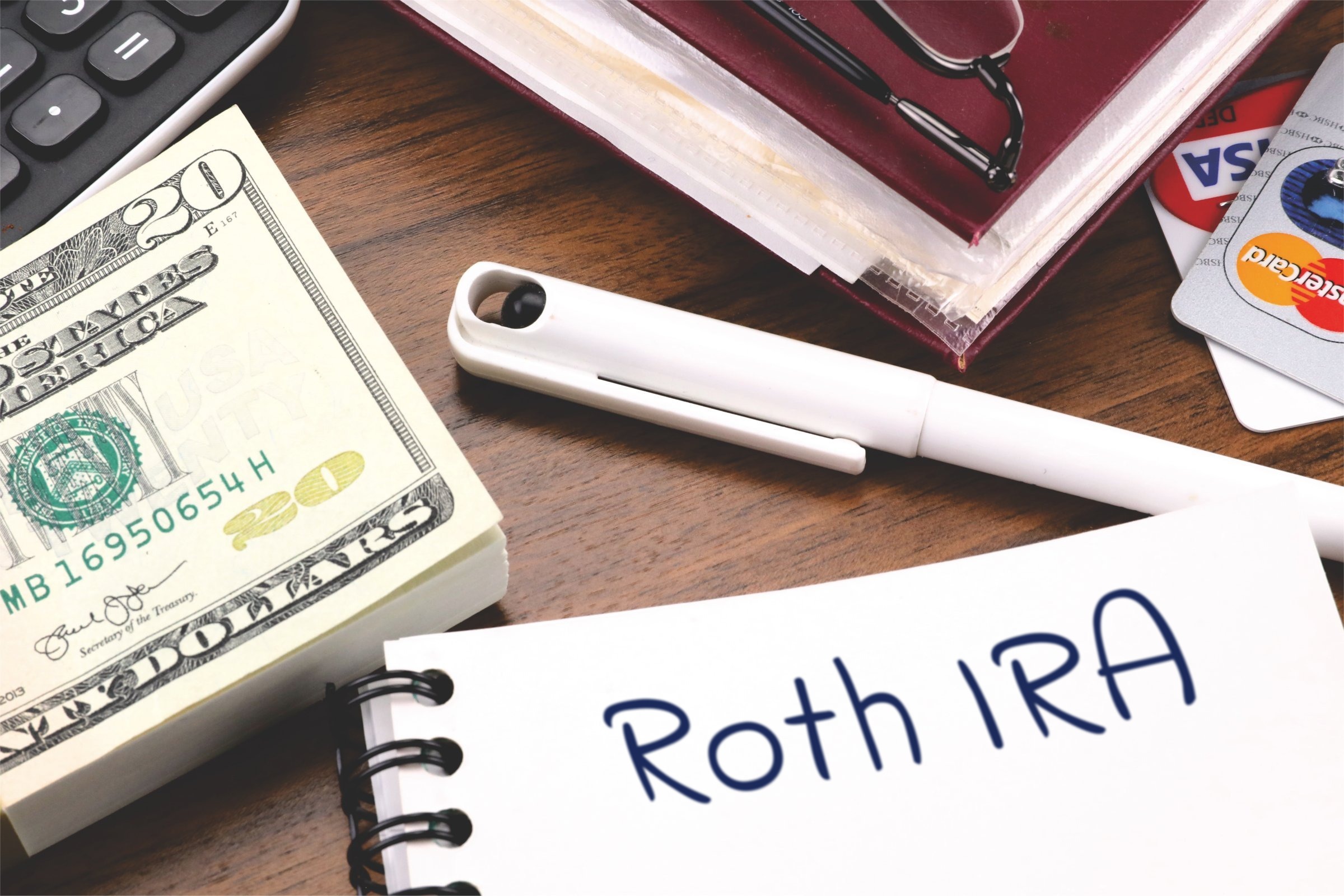 Spiral notepad with a blank page, pen and dollar bills template with the words "Roth IRA" printed on the surface
