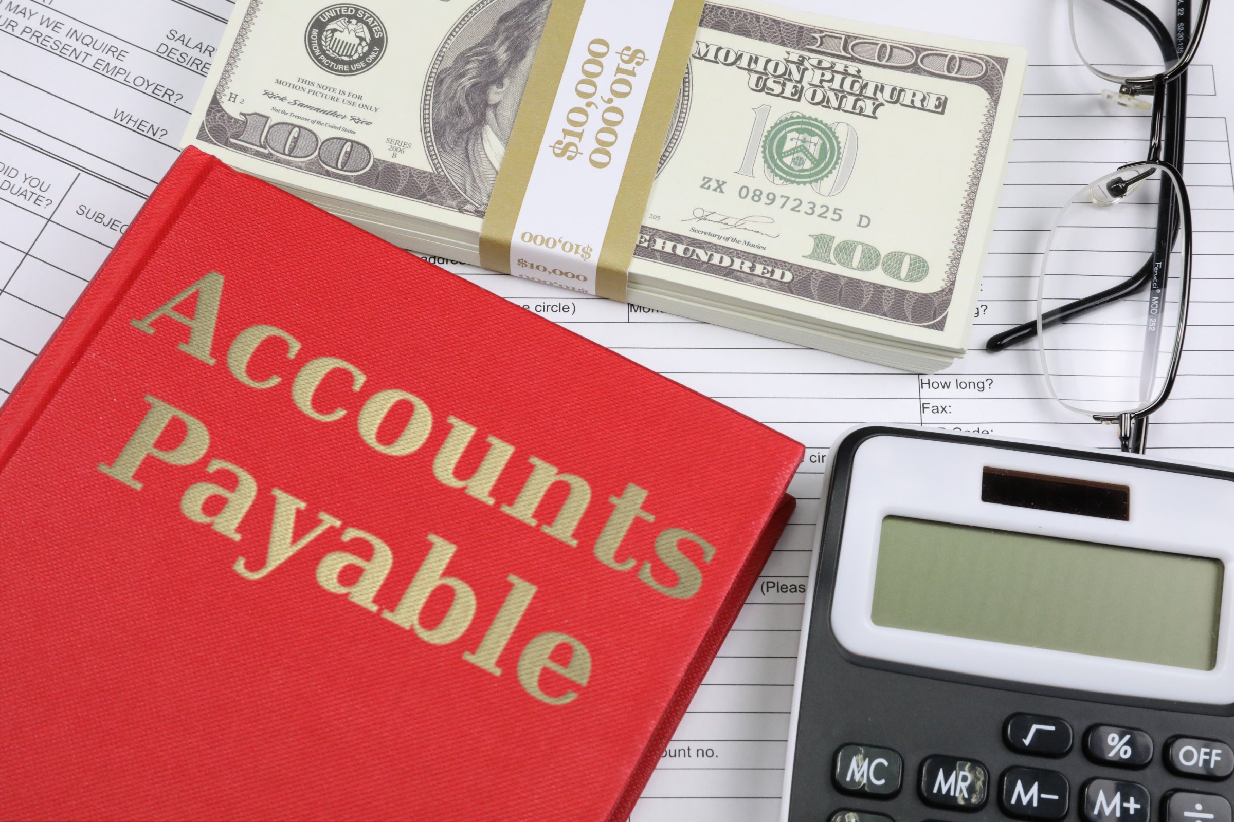 Red hardback book with dollars, glasses and calculator template with the words "Accounts Payable" in metallic foil lettering