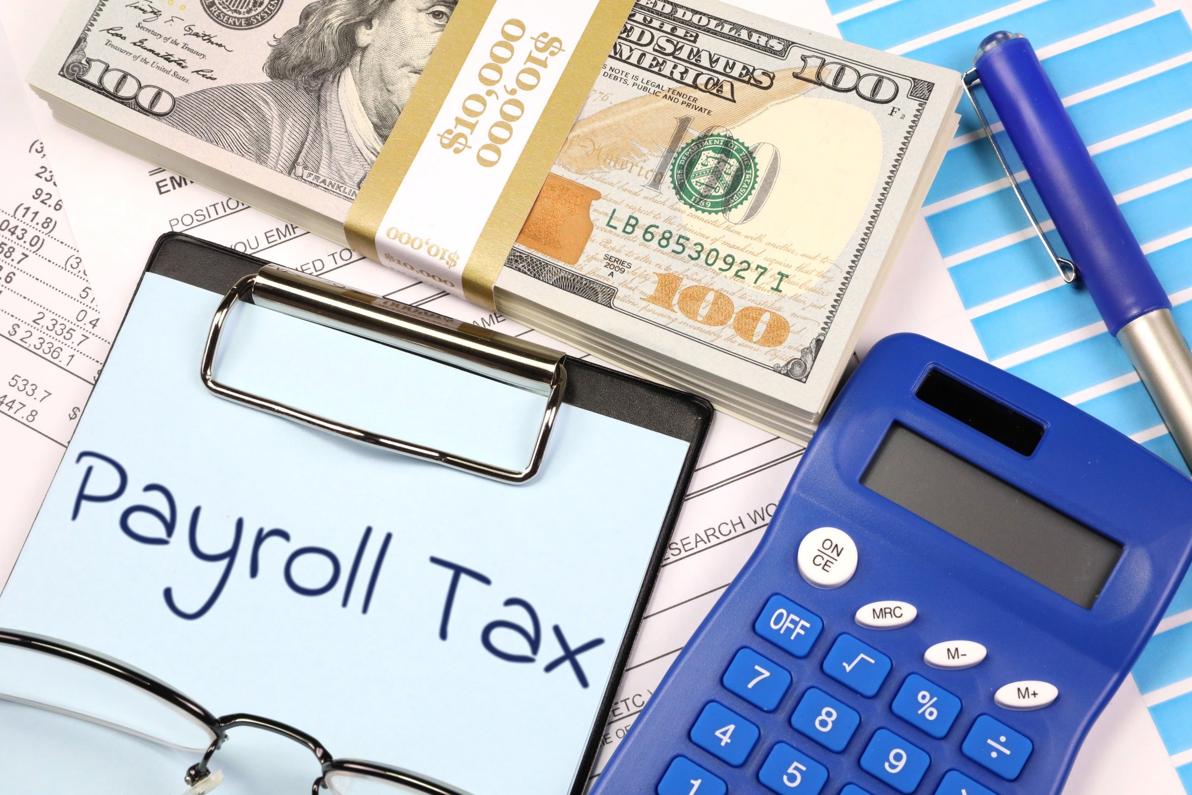 Clipboard with blue paper, dollars and calculator template with the words "Payroll Tax" printed on the surface