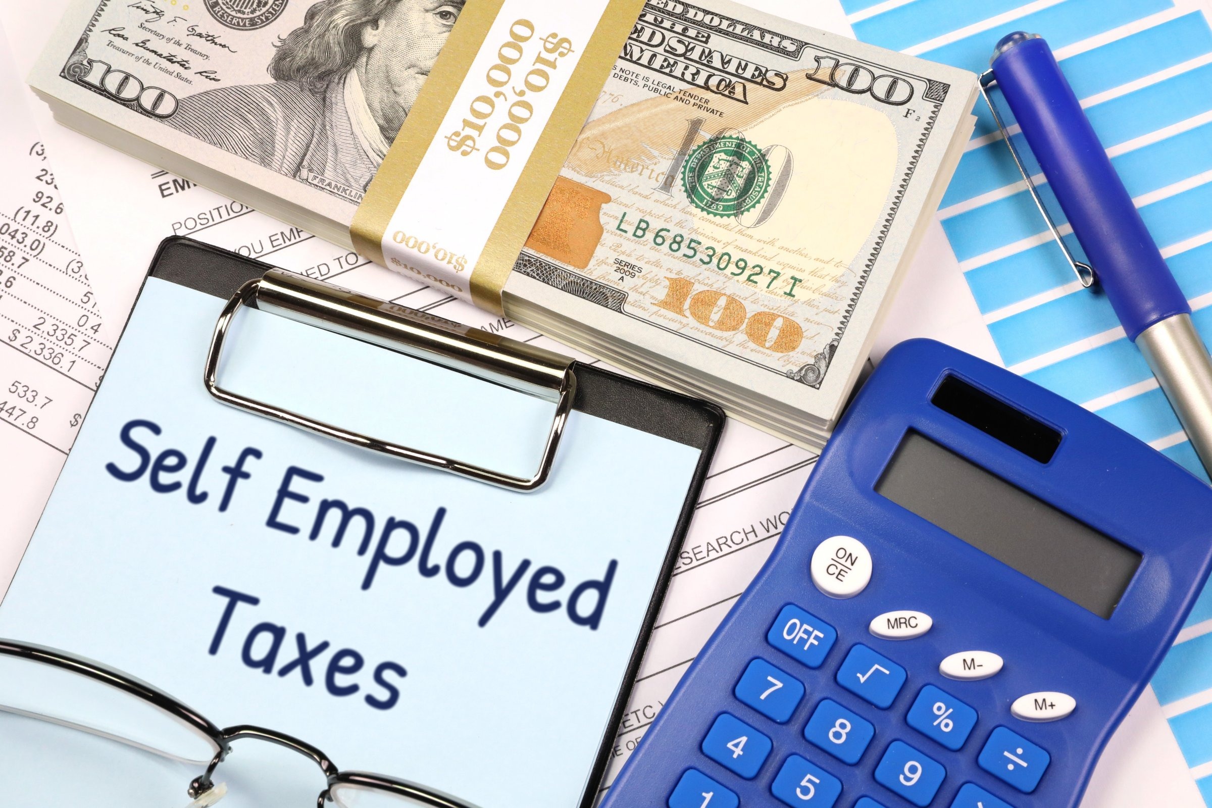 Clipboard with blue paper, dollars and calculator template with the words "Self Employed Taxes" printed on the surface
