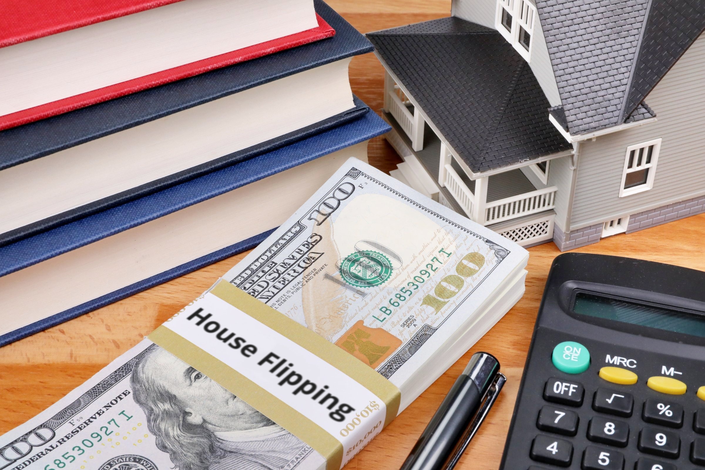 Stack of dollars with a banded label, model house and books template with the words "House Flipping" printed on the surface